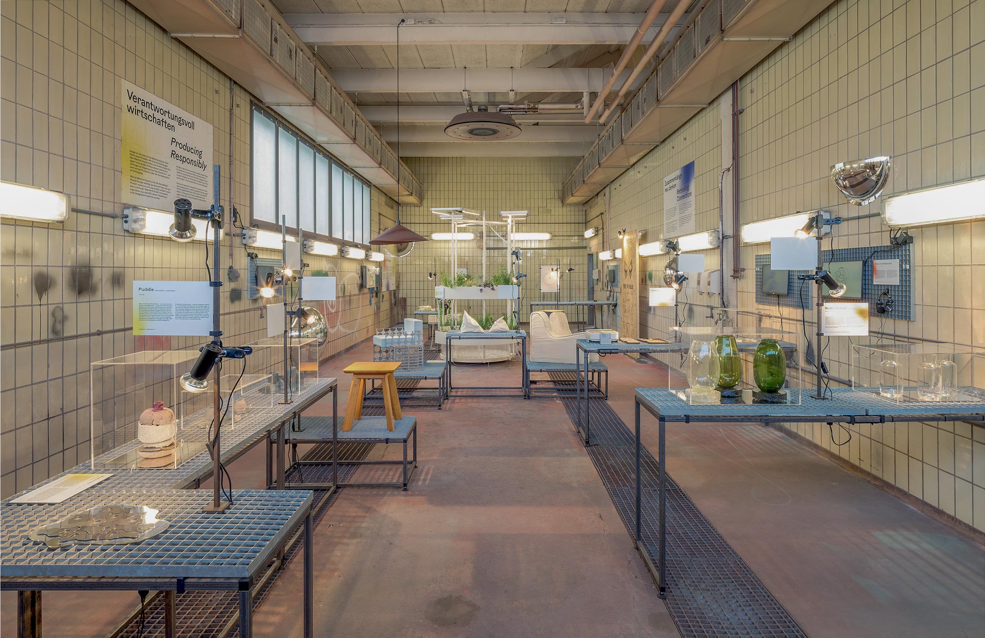 Industrial-style exhibition room with various objects on metal tables, illuminated by lamps.