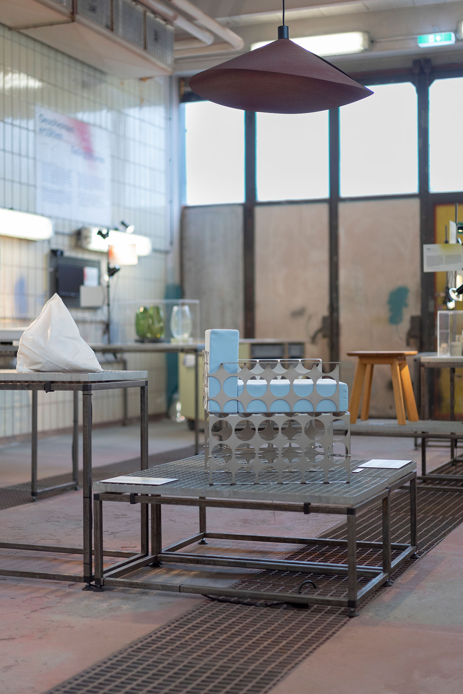 Industrial-style exhibition room with a aluminium chair on a metal plinth, illuminated by lamps.